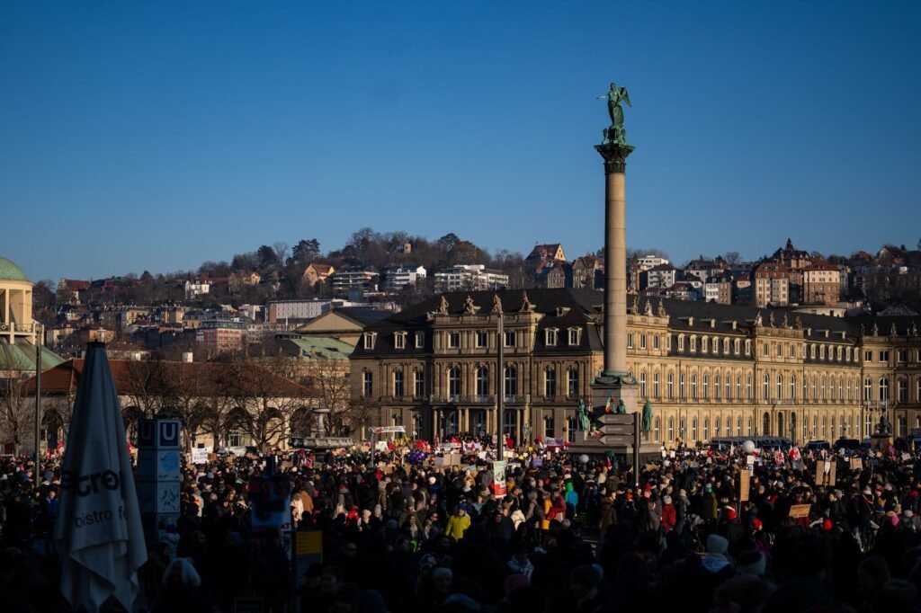 Eine große Menschenmenge versammelt sich auf der Schlossplatz in Stuttgart, mit historischen Gebäuden und Wahrzeichen im Hint